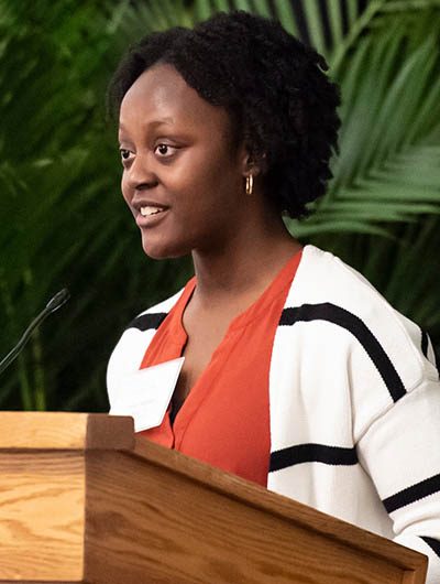 Fodun Ologunde in an orange shirt and black and white jacket stands at a podium