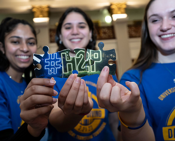 Three people hold up puzzle pieces that combine to read #H2P