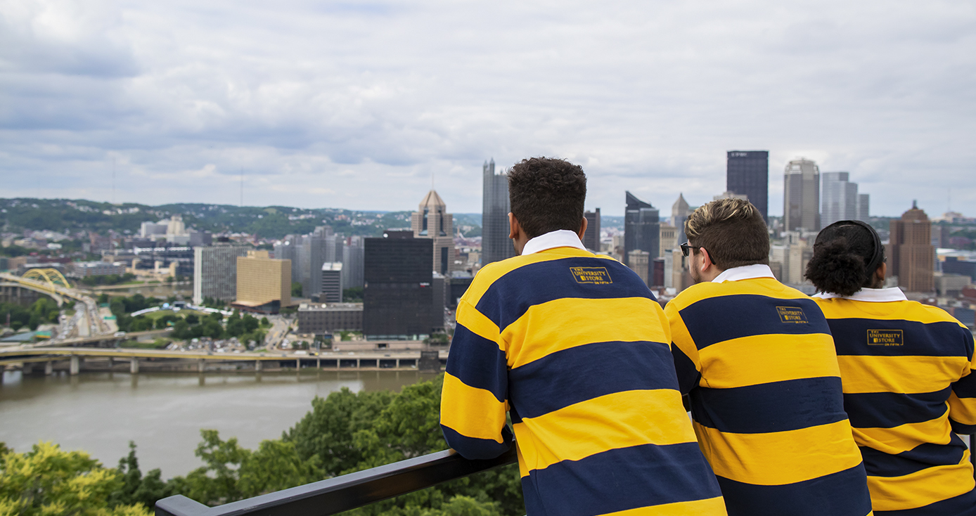 Three Pitt Pathfinder students wearing blue and gold striped rugby shirts look out over the city of Pittsburgh skyline