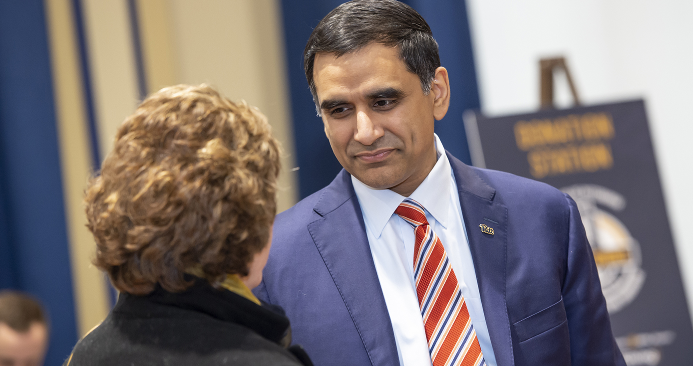 A man in a blue suit with a small pin on his lapel and a red striped tie speaks with another person