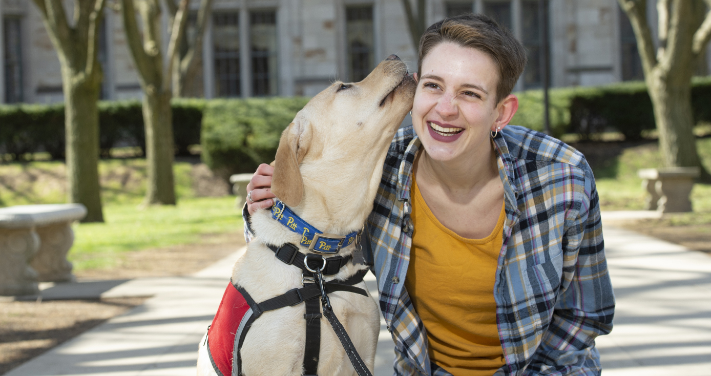 Emma Oaks wearing plaid shirt over yellow t-shirt being licked on the face by a service dog in training