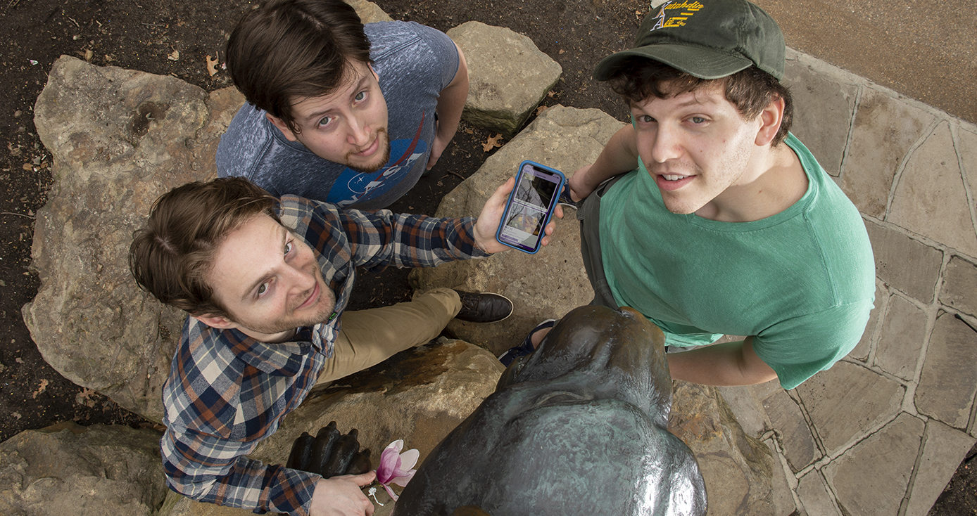 three young men standing next to the Panther statue on campus, looking up. One is holding a phone with the app on the screen