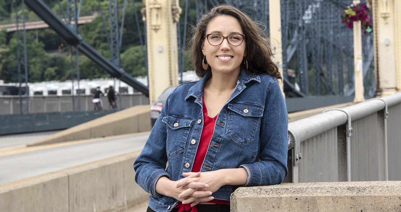 Jenna Baron standing on a walkway on a bridge in the city of Pittsburgh. Wearing denim jacket and red shirt. The bridge is painted yellow.