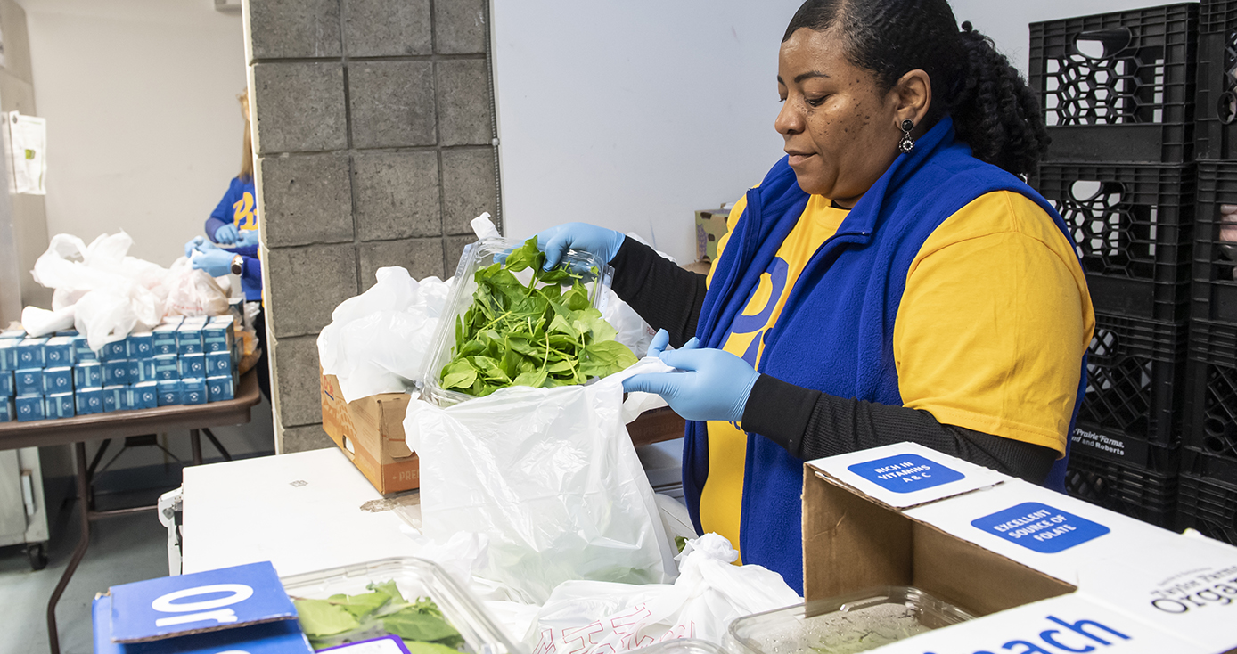 A woman in a yellow shirt and blue vest packs vegetables into a plastic bag