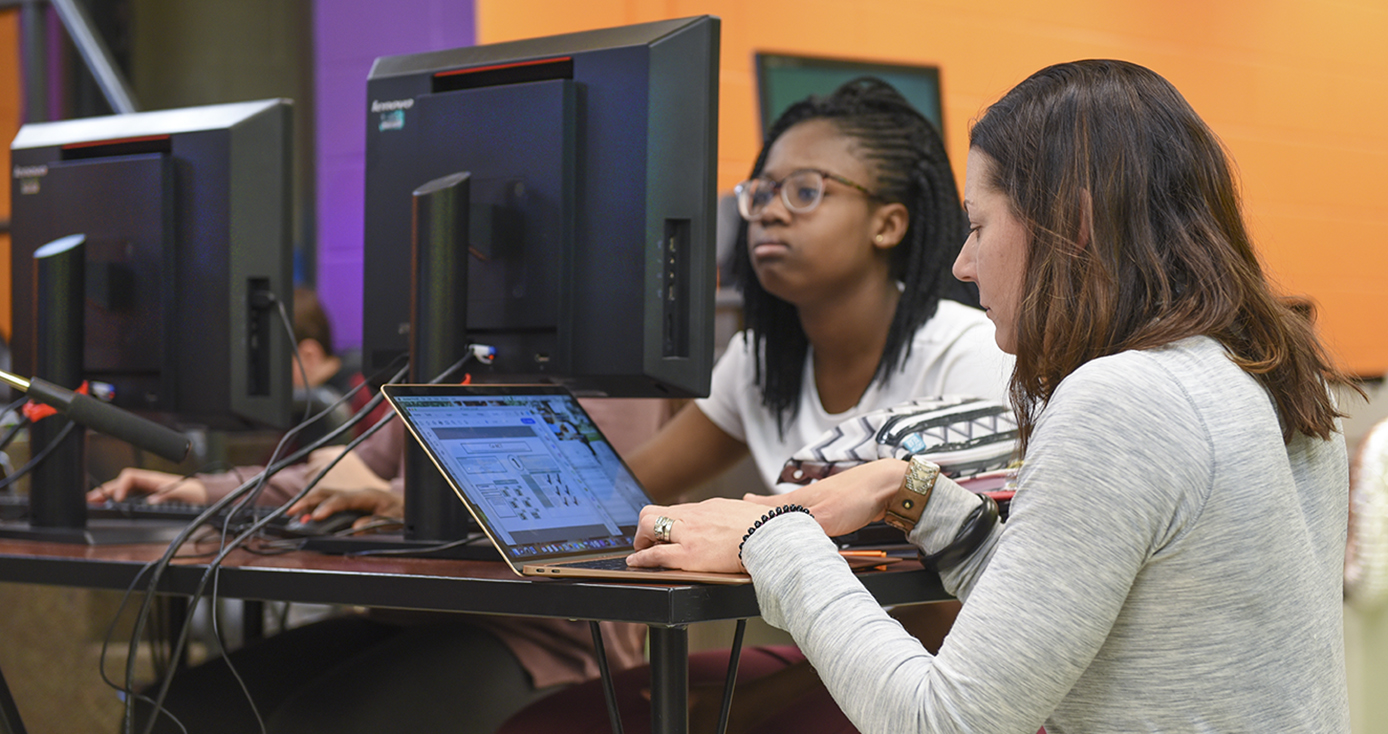 a teacher at a laptop and a student at a desktop, both working