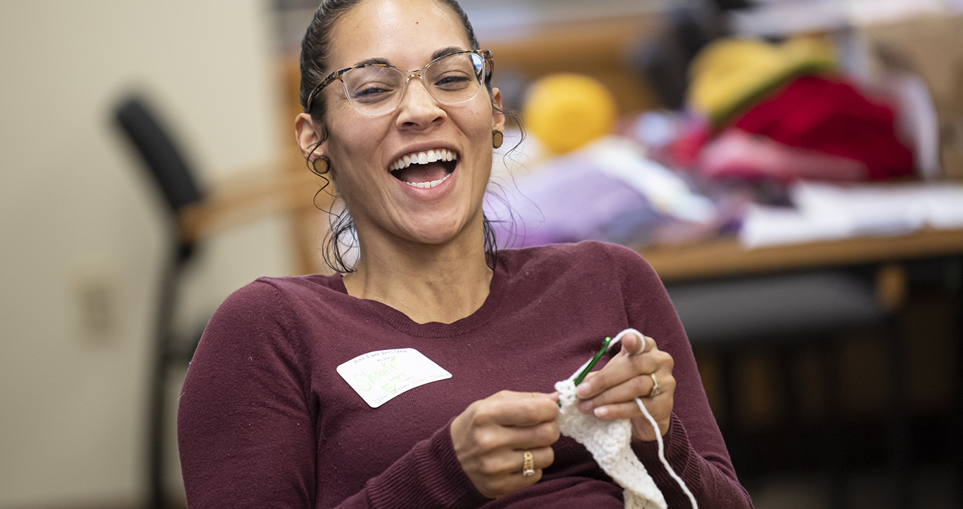 A woman in a maroon long sleeve shirt laughs while knitting