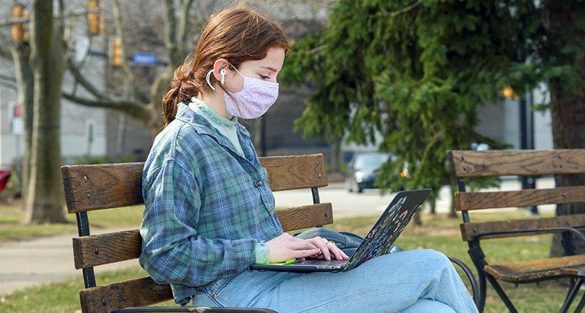 A person in a plaid shirt and pink face mask sits on a bench with a laptop