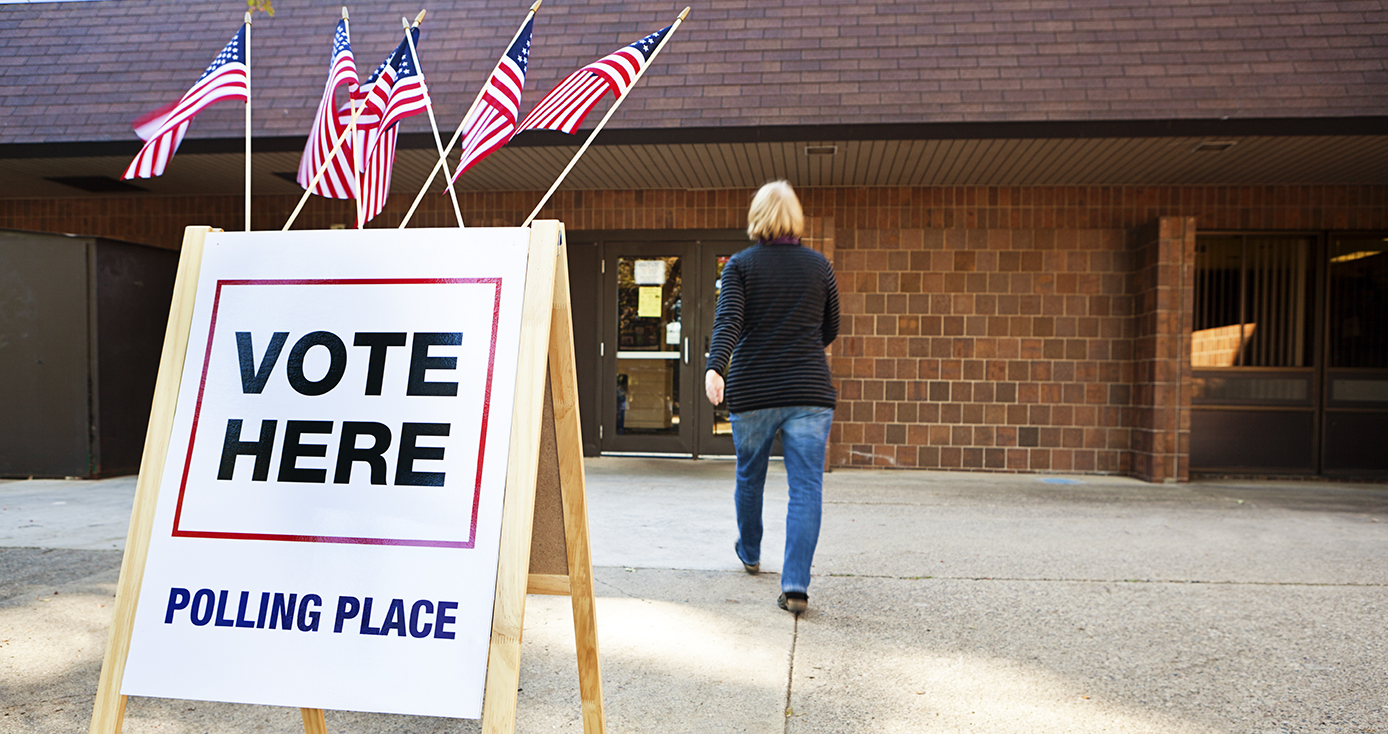 Brick building in background with person walking toward it a "Vote Here' sign in the foreground with American flags on top of it