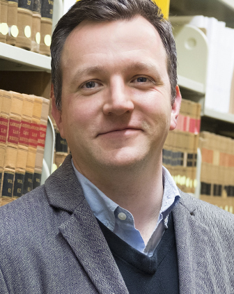 Morgan in a collared shirt, vest and jacket in front of a bookcase