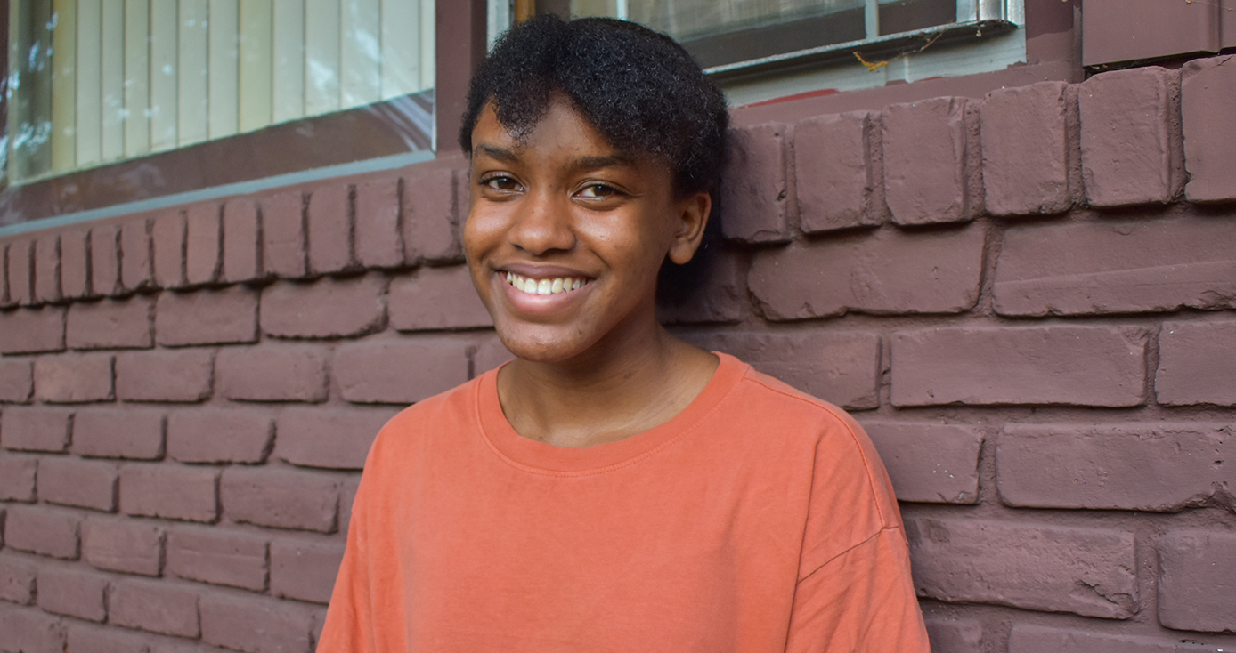 A woman in an orange shirt leaning against a brick wall