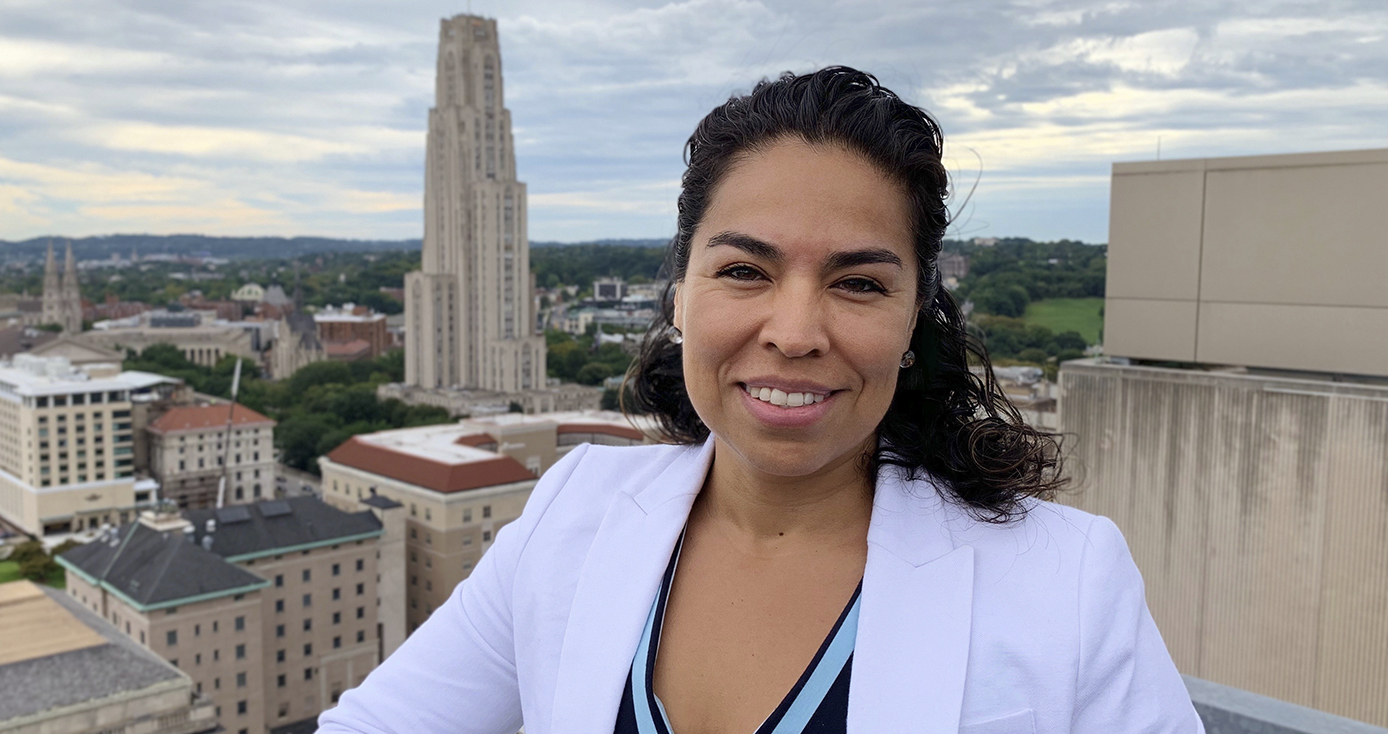 Gelsy Torres-Oviedo in white jacket over blue shirt, standing on a rooftop overlooking Pitt campus with Cathedral of Learning prominently in the background