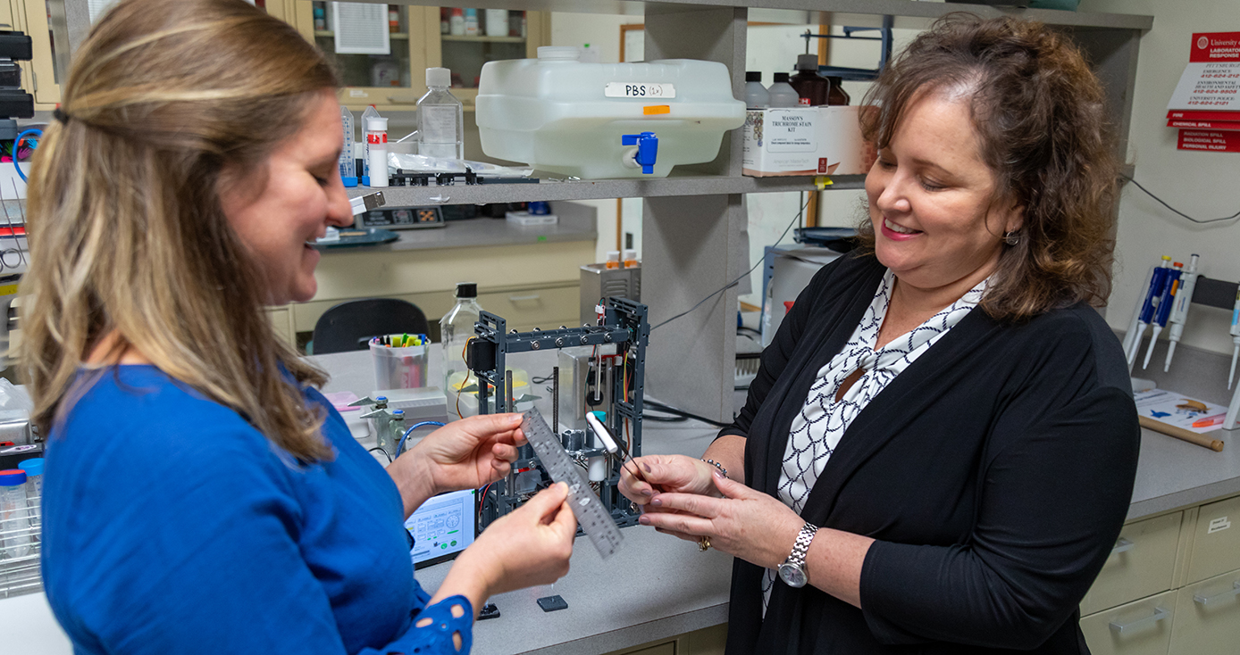 Two women stand in a lab, looking down at equipment and smiling