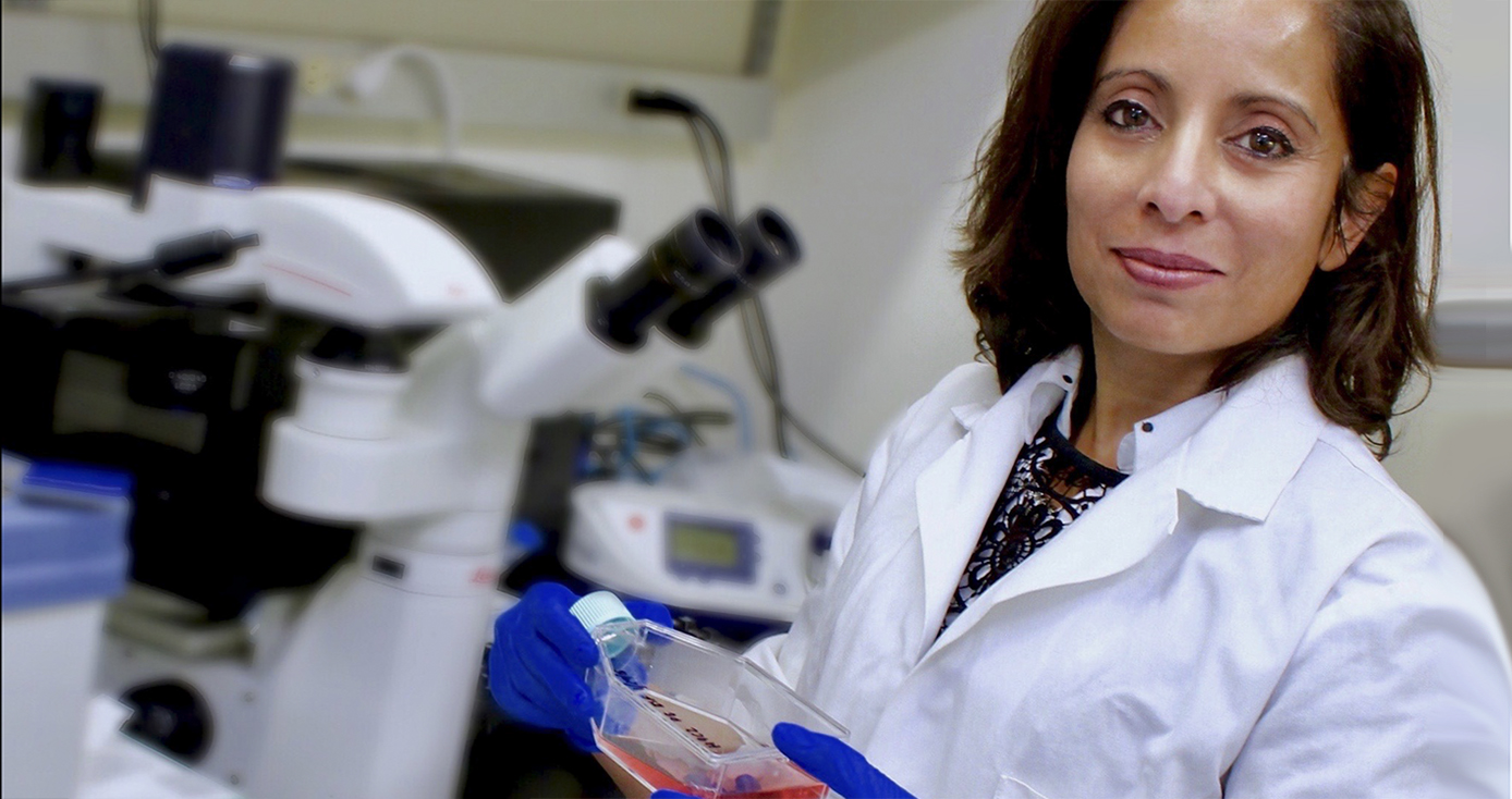 Maliha Zahid in white lab coat and wearing blue gloves holding a clear bottle of liquid in a lab setting