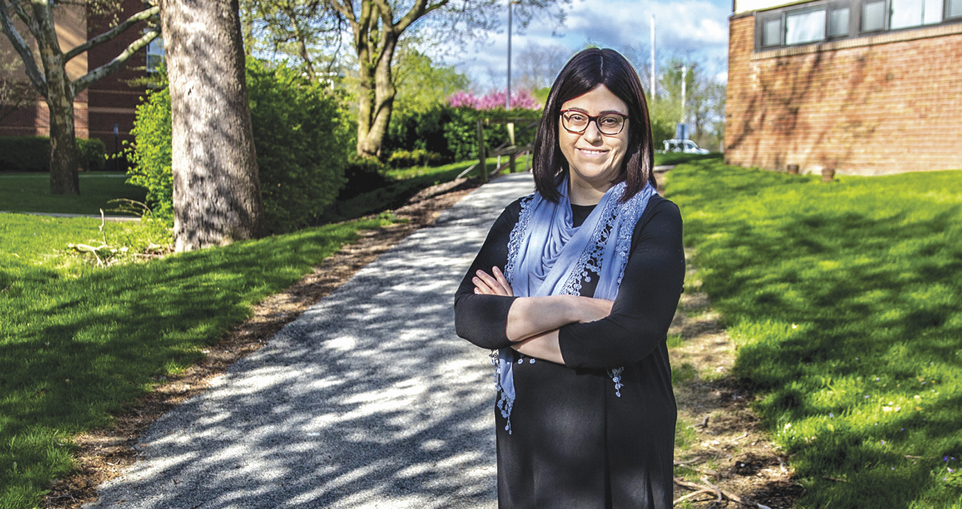 Massart standing in a green space between a couple of brick buildings. She's wearing a black dress with three-quarter-length arms and a blue shawl, and has her arms crossed over her chest.