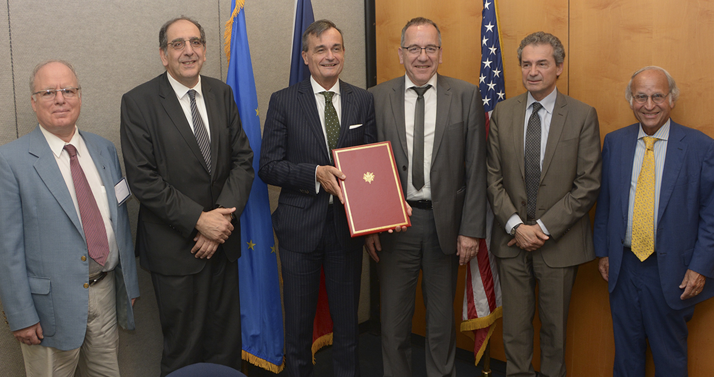 six men in suits standing in front of three flags; the man three from the left is holding a red bound version of the agreement