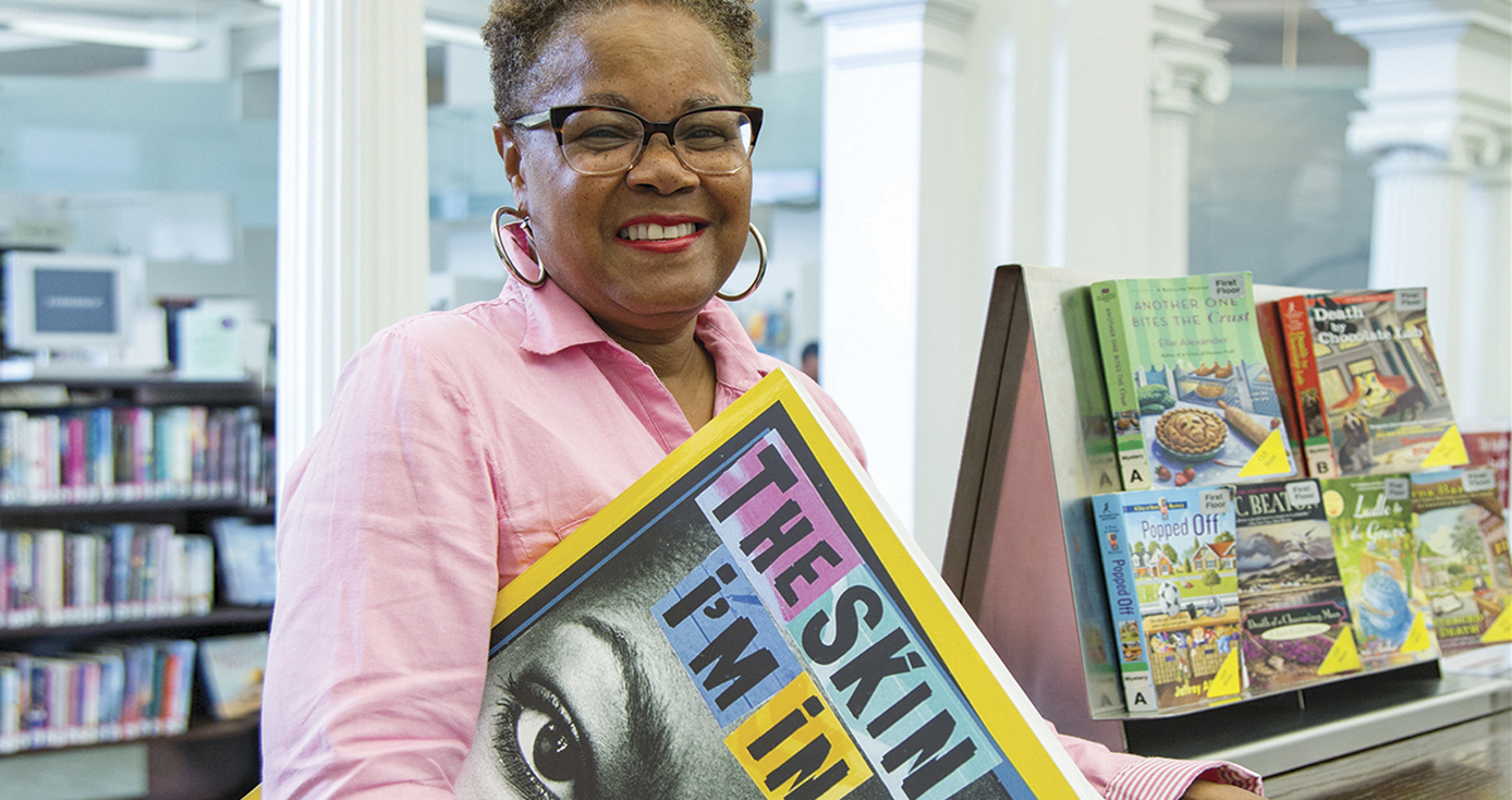 Sharon Flake, holding oversized copy of her book "The Skin I'm In," standing in a library with shelves of books in the background