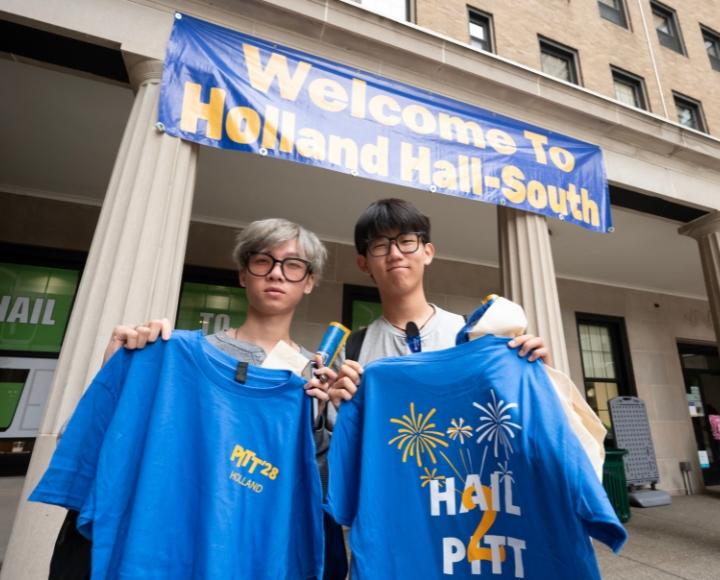 Two people hold up shirts in front of a Holland Hall banner