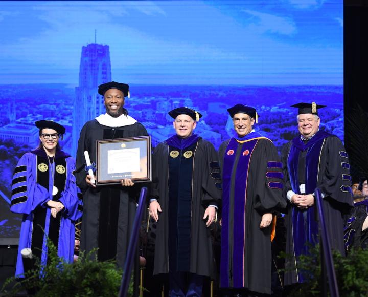 Five people wearing graduation regalia stand on stage at commencement.