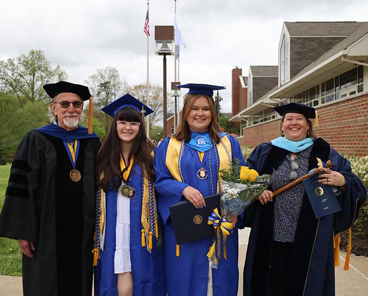 Two graduates stand in between two faculty members, all in regalia.