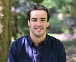 Man with dark hair sits in a wooded area and smiles for camera.