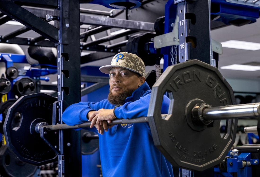 Man wearing a baseball cap leans on free weights in a weight room.