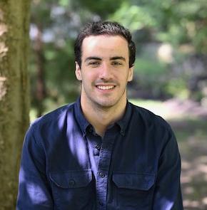 Man with dark hair sits in wooded area and smiles for camera.