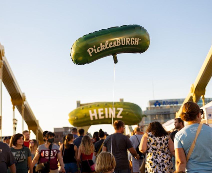 Many people on one of the Pittsburgh bridges, a child carries a pickle-shaped balloon that reads "PicklesBURGH". In the distance there is a pickle-shaped sign reading "HEINZ".