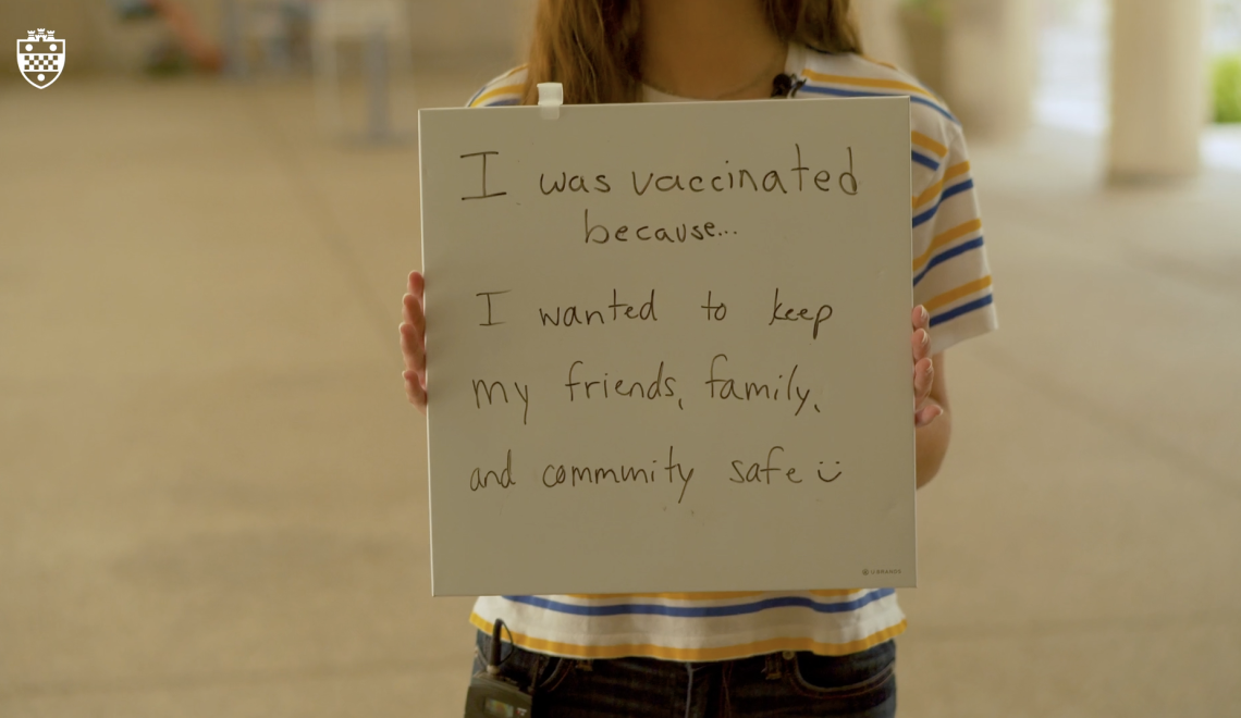 a person in a white, blue and gold shirt holding up a sign that says "I was vaccinated because...I wanted to keep my friends, family, and community safe :)"